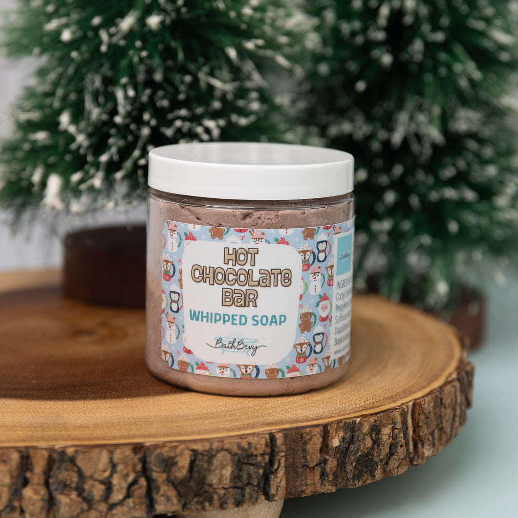 A container of HOT CHOCOLATE BAR WHIPPED SOAP by Bath Bevy, placed on a wooden surface with a decorative pattern and Christmas tree branches in the background.