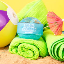 Load image into Gallery viewer, Bath bomb with a blue container on green towels and a beach ball, against a yellow background
