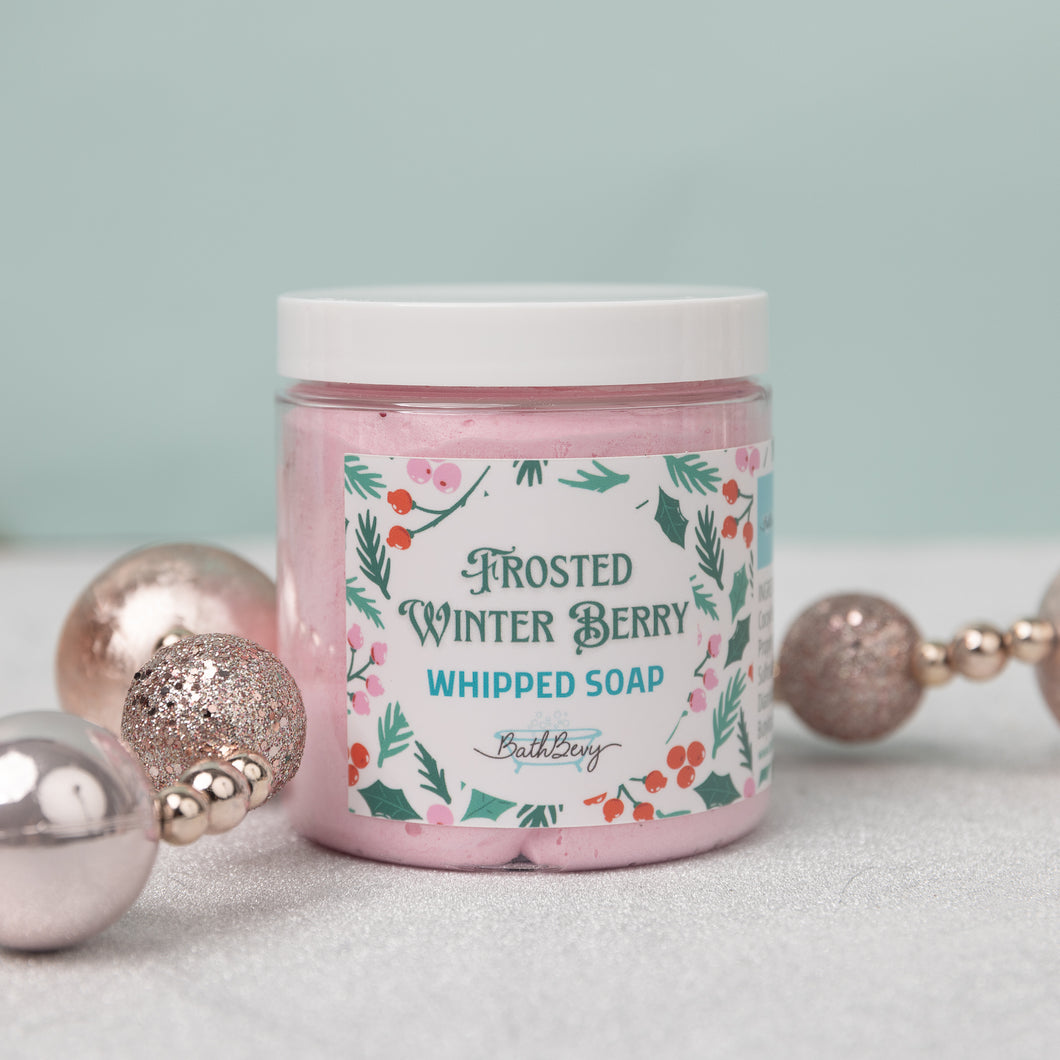 A jar of Frosted Winter Berry Whipped Soap by Bath Bevy, decorated with holiday ornaments on a light background.