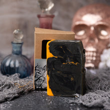 Load image into Gallery viewer, A black handmade soap bar with yellow accents, placed on a cardboard box that has &#39;ALCHEMY&#39; written on it. The box is labeled &#39;DIY Soap Bar&#39; and has a skull design in the background.