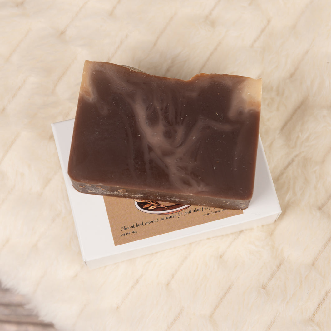 A brown handmade soap bar resting on a white dish, with a fuzzy white surface in the background.