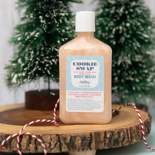 A bottle of Cookie Swap body wash by Burt's Bees, pump bottle design, with a label that reads 'holiday'. The bottle is placed on a wooden surface with a decorative backdrop of Christmas trees and red and white twine.