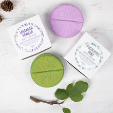 Load image into Gallery viewer, Two bath bombs, one purple and one green, with their packaging on a light wooden surface.